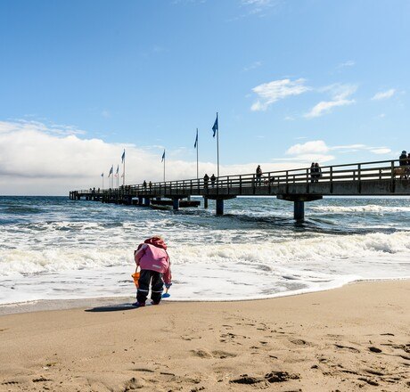 Kind spielt am Strand bei Wind und wetterfest, im Hintergrund die Seebrücke