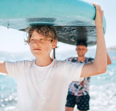Blonde teenage son with father surfers smiling carrying the stand-up paddleboard with a paddle on the bright sunny day noon. Active family summer vacation time near the sea concept image.