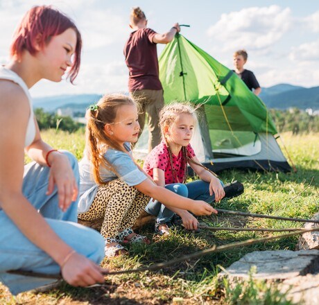 Three sisters smiling and roasting marshmallows and candies on sticks over a campfire flame while two brothers set up the green tent. Happy family outdoor picnic camping activities concept.