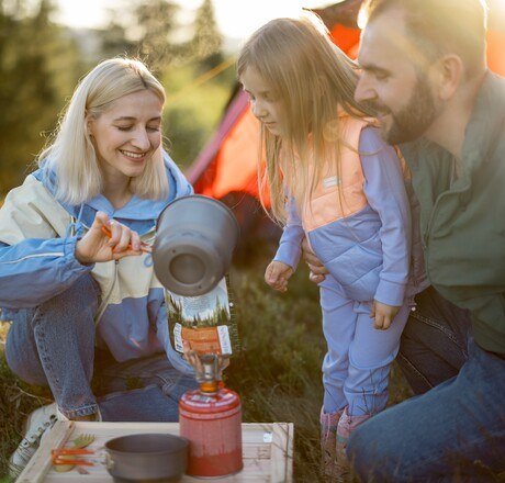 Young couple with little girl cooking dried-food for hiking, pouring boiling water into the package. Family at campsite making food for travelers. Sublimated food and family camping concept